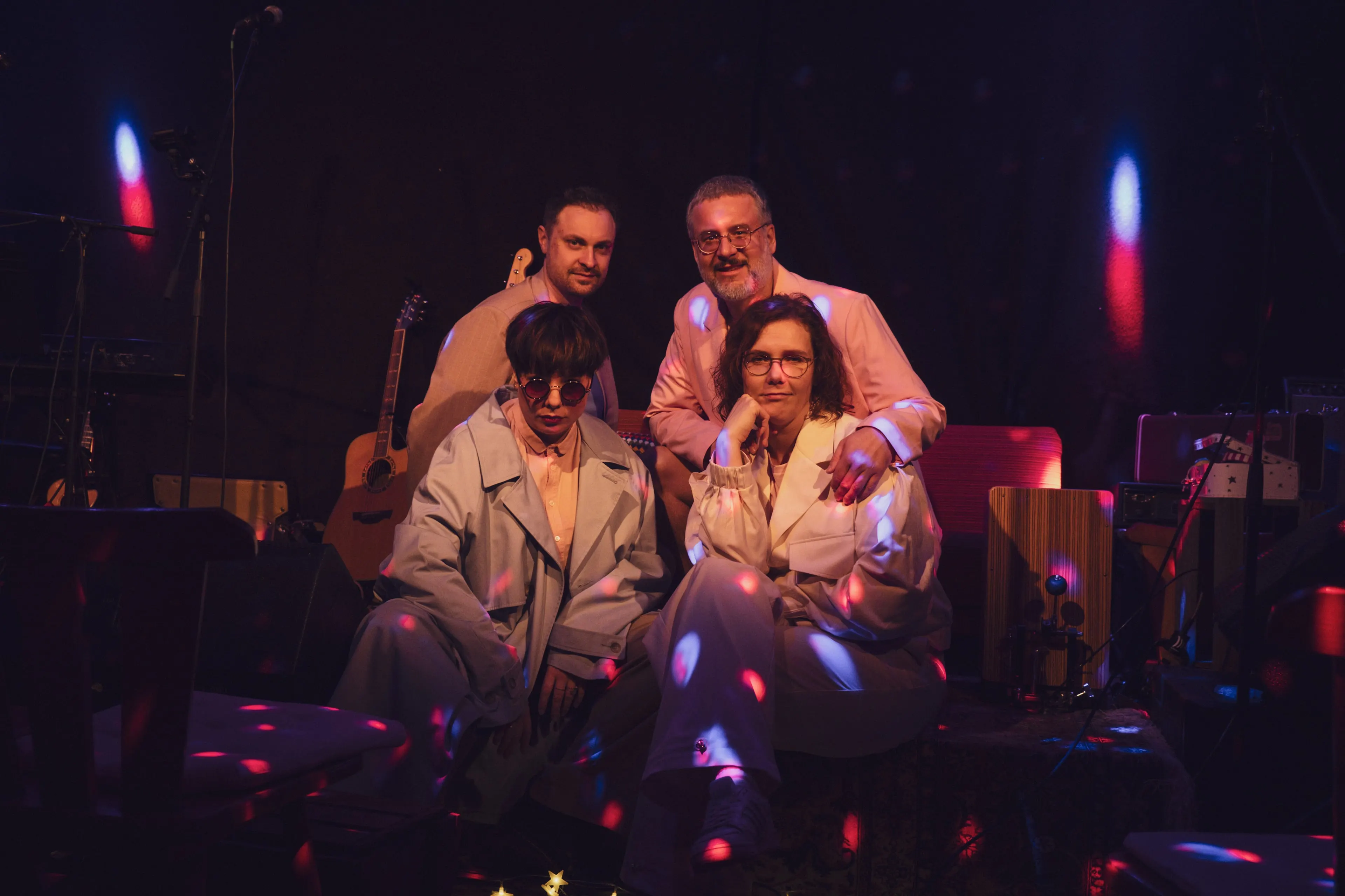 Die vier Bandmitglieder auf einer abgedunkelten Bühne, in hellen Anzügen und dicht beieinander, dahinter eine Akustikgitarre und weitere Instrumente, im Raum farbige Lichtpunkte.